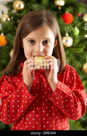 Young Girl Eating Star Shaped Christmas Cookie in front of Christmas Tree Banque D'Images