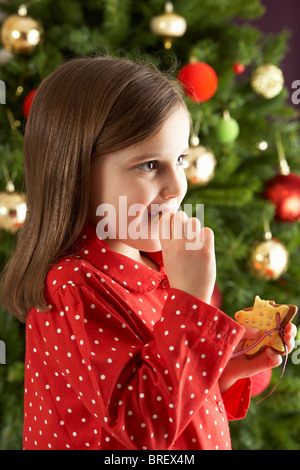 Young Girl Eating Star Shaped Christmas Cookie in front of Christmas Tree Banque D'Images