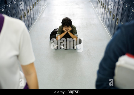 Teenage boy sitting sullenly sur le plancher du couloir d'école, les camarades de classe en premier plan Banque D'Images