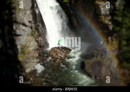 Un jeune homme se tient à la base d'une cascade à côté d'un arc-en-ciel avec ses bras ouverts. Banque D'Images