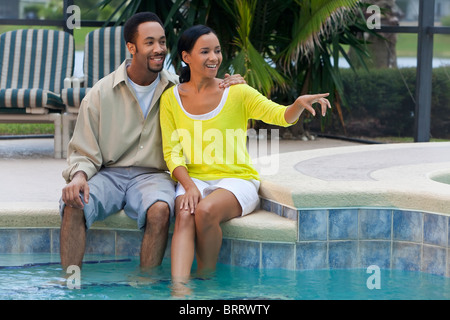 Une happy African American man and woman couple dans la trentaine assis à l'extérieur par un pool de sourire et de pointing Banque D'Images