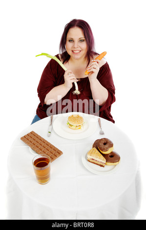 Young, fat woman holding vegetables devant une table avec des hamburgers et des gâteaux Banque D'Images