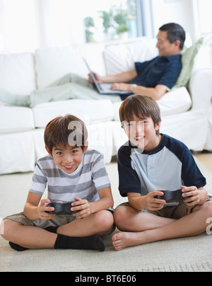 Smiling frères jumeaux jeux vidéos tandis que le père utilise un ordinateur portable sur salon canapé Banque D'Images