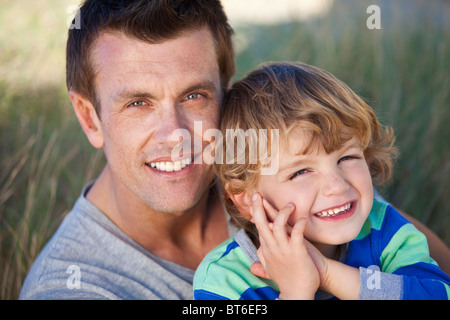 Un homme et jeune garçon, père et fils, s'asseoir et s'amuser dans l'herbe des dunes de sable d'une plage ensoleillée Banque D'Images