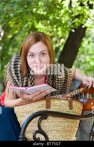 Red-haired woman assise sur un banc avec réserve et souriant à l'appareil photo Banque D'Images