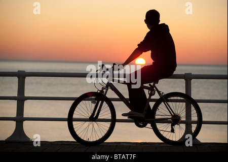Soirée octobre - Silhouette d'un garçon sur un vélo qui se profile au coucher du soleil sur la baie de Cardigan, Wales Aberystwyth UK Banque D'Images