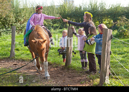 Little girl lying équitation un poney, d'autres pour la regarder et lui donner quelque chose Banque D'Images