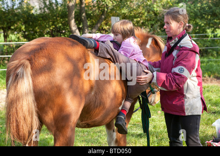 Petite fille grimper sur un poney avec l'aide de son professeur d'équitation Banque D'Images