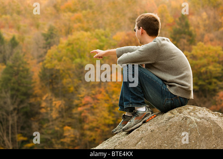 Young male hiker en jeans pointant tout en restant assis sur un rocher en face de feuilles d'automne Banque D'Images