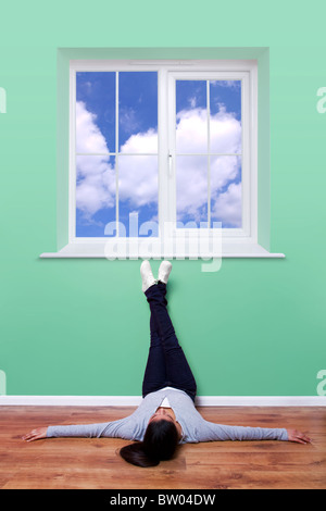 Jeune femme allongée sur le sol avec ses pieds vers le haut du mur de penser comme elle regarde par la fenêtre pour un ciel bleu clair. Banque D'Images