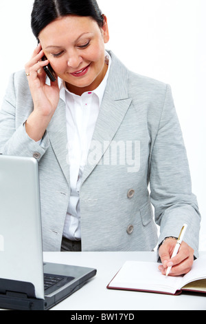 Secrétaire générale d'occupation sur le téléphone et la prise de notes dans le bloc-notes avec un ordinateur portable à proximité Banque D'Images