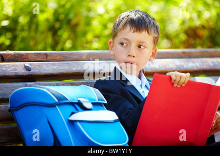 Portrait de smart garçon assis dans le parc avec livre intéressant et à côté Banque D'Images