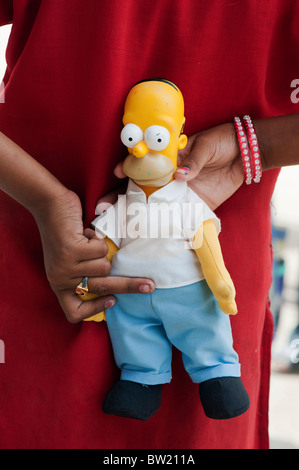 Jeune Indien street girl holding a Homer Simpson doll derrière son dos. L'Andhra Pradesh, Inde Banque D'Images