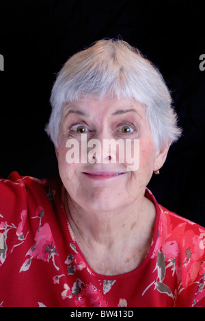 Une femme âgée aux cheveux blancs portant une chemise fleurie rouge fait une drôle de face avec un sourire niais et de grands yeux. Banque D'Images