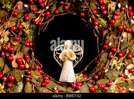 Raphia décoratif ange de Noël à l'intérieur d'une couronne de Noël avec des feuilles vertes et des fruits rouges. Sur noir. Banque D'Images