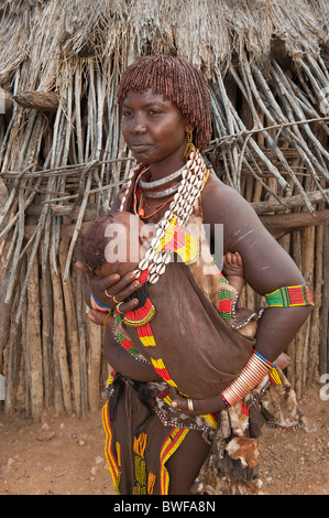 Jeune femme Hamar avec argile rouge dans ses cheveux et de colliers faits de cauris et tenant son bébé dans les bras, l'Ethiopie Banque D'Images