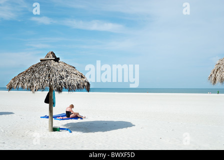 Femme lisant sur plage de sable blanc avec de la paille à proximité parapluie abri soleil ombre coulée. Banque D'Images