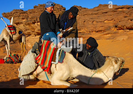 Les nomades touaregs aider un touriste femelle à monter sur un dromadaire dans le désert du Sahara, montagnes Acacous, Libye Banque D'Images