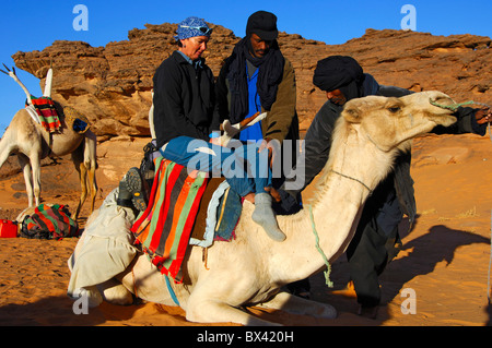 Les nomades touaregs aider un touriste femelle à monter sur un dromadaire dans le désert du Sahara, montagnes Acacous, Libye Banque D'Images