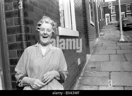 Riant, une femme blanche de la classe ouvrière des années 1980 dans des bigoudis portant sa maison de ménage debout devant sa maison mitoyenne riant Blackburn Lancashire Angleterre 1983 UK HOMER SYKES Banque D'Images