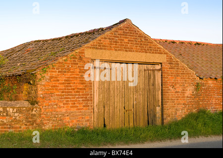 Ancienne grange en brique rouge avec porte en bois face à Road Banque D'Images