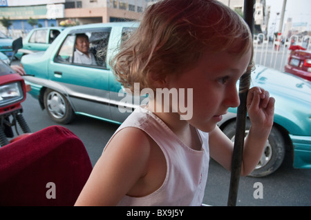 Petite fille blonde à l'intérieur d'un excès de rouge tricycle le long d'un boulevard animé à Datong, Shanxi, en Chine. Banque D'Images