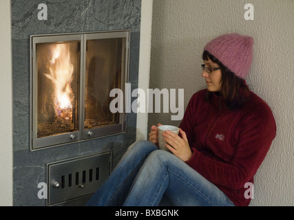 Une jeune femme assise par une cheminée tenant une tasse de café. Banque D'Images