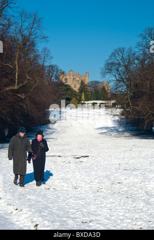 Un vieux couple marchant dans la neige au parc de Wollaton, Nottingham, Angleterre, Royaume-Uni. Avec Wollaton Hall dans l'arrière-plan. Banque D'Images