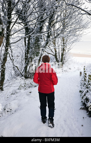 Le Nord du Pays de Galles, Royaume-Uni. Walker en veste rouge sur la neige couverts Isle of Anglesey Sentier du littoral menant à la plage en hiver Banque D'Images