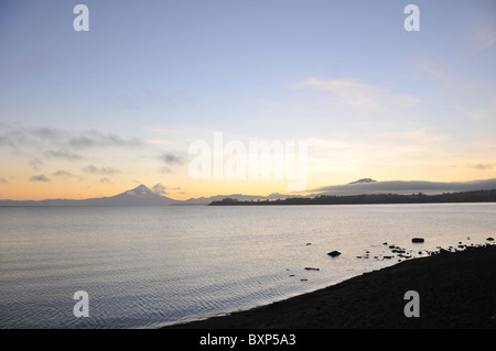 Le lac Llanquihue vue, d'une obscurité Puerto Varas plage, d'une aube du ciel soleil jaune glow sur les volcans Osorno et Calbuco, Chili Banque D'Images