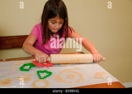5-year-old fille blanc/philippine de rouleaux sur la pâte avec un rouleau à pâtisserie sur un papier ciré à la table de cuisine Banque D'Images