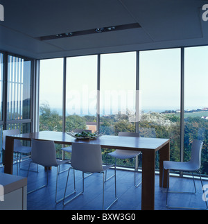 Table en bois rectangulaire et chaises blanches avec pieds métalliques dans une salle à manger moderne avec vue sur l'océan par de grandes fenêtres Banque D'Images