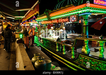 ISTANBUL, Turquie — des bateaux de pêche éclairés servent des dîners le long du front de mer d'Eminonu, proposant des balik ekmek (sandwichs de poisson) traditionnels dans leurs cuisines flottantes. Les bateaux éclairés créent une scène gastronomique nocturne distinctive le long du front de mer historique. Les clients assis à des tables au bord de l'eau dégustent du poisson grillé frais sous les lumières du soir. Banque D'Images