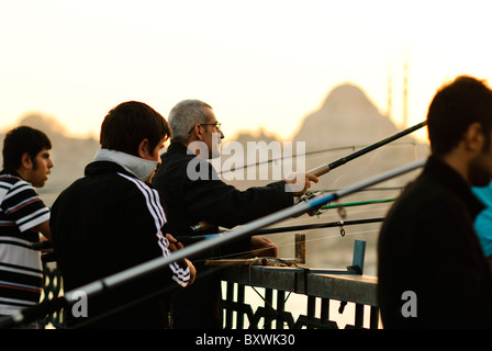 ISTANBUL, Turquie — les pêcheurs bordent les balustrades du pont historique de Galata, jetant leurs lignes dans la Corne d'Or ci-dessous. Le pont à deux niveaux, qui relie Eminonu à Karakoy, accueille des véhicules, des tramways et des piétons sur son pont supérieur et abrite des restaurants sur son niveau inférieur. La silhouette distinctive de la mosquée Suleymaniye du XVIe siècle domine l'horizon au-dessus du pont. Banque D'Images