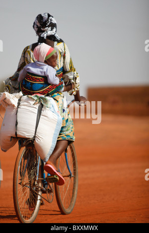 À Djibo dans le nord du Burkina Faso, une femme porte son jeune enfant attaché sur son dos alors qu'elle chevauche à travers la ville sur un vélo. Banque D'Images