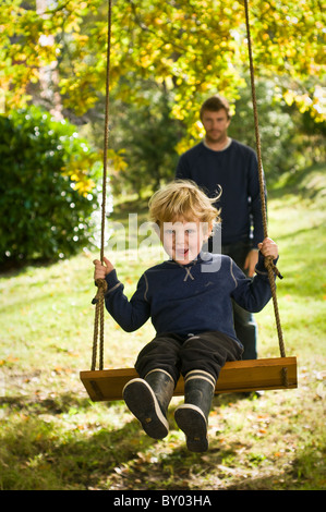 Jeune garçon jouant sur une balançoire dans un jardin, qui est poussé par son père. Banque D'Images