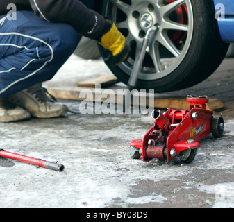 Un cric rouleur hydraulique rouge à côté d'une voiture tandis qu'un mécanicien est de dévisser les écrous de roue avec une clé pour démonter la roue. Banque D'Images