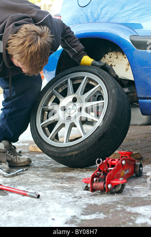 Un jeune homme de race blanche mechanic dépose d'une roue d'une voiture, qui s'est tenue en place par un cric sur une allée glacée. Banque D'Images