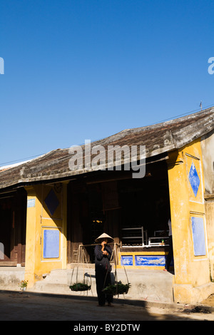 Vieille dame transportant des légumes en face d'un bâtiment historique de Hoi An, Vietnam Banque D'Images