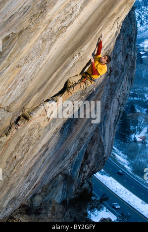 Un alpiniste dans une chemise rouge et jaune atteignant jusqu'à une prise sur une route escarpée et difficile. Banque D'Images