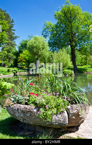 Un semoir en pierre avec des géraniums en face d'un grand bassin d'agrément ou d'un petit lac dans un jardin de campagne anglaise en été Banque D'Images