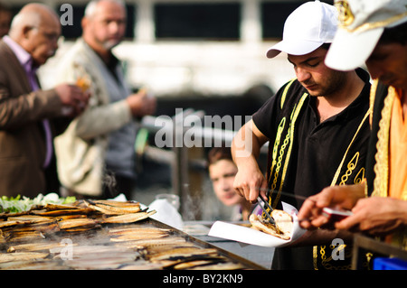 Balik Ekmek Fish Sandwich Street Food Istanbul Turquie // ISTANBUL, Turquie — Un vendeur de rue grillera du poisson frais sur le front de mer d'Eminonu, près du pont de Galata, préparant le traditionnel balik ekmek (sandwich au poisson). Le balik ekmek, généralement préparé avec du maquereau grillé servi dans du pain blanc avec des oignons et de la laitue, est l'un des plats de rue les plus populaires d'Istanbul. Les vendeurs de poissons riverains opèrent à partir de bateaux flottants et de stands le long de la Corne d'Or à cet endroit depuis des générations. Le quartier d'Eminonu sert de terminal de ferry majeur et de plaque tournante commerciale reliant l'Europe et l'Asie d'Istanbul Banque D'Images