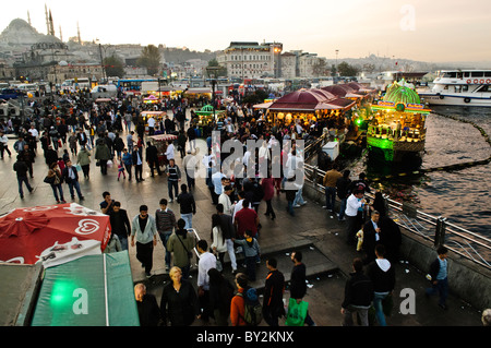 Balik Ekmek sandwichs de poisson Eminonu Waterfront Istanbul // ISTANBUL, Turquie — des bateaux de pêche éclairés servent des balik ekmek (sandwichs de poisson) traditionnels le long du front de mer animé d'Eminonu, au pied du pont de Galata. La scène du soir capture la culture culinaire dynamique du front de mer avec des mosquées historiques silhouettes sur le ciel assombrissant. Les vendeurs locaux perpétuent une tradition séculaire de servir du poisson grillé frais à partir de leurs bateaux. Banque D'Images