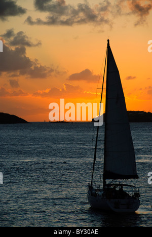 ÎLES VIERGES AMÉRICAINES — Un voilier solitaire glisse paisiblement à travers des eaux calmes vers un spectaculaire coucher de soleil des Caraïbes dans les îles Vierges américaines. La silhouette du bateau est dramatiquement contrastée avec les oranges vibrantes, les roses et les violets peignant le ciel du soir. Banque D'Images