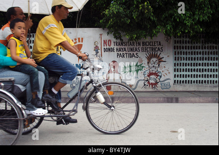 Scène de rue à Holguin (Cuba). Un bicitaxi équitation avec les clients et les graffitis sur le mur en arrière-plan la prévention des vélos. Banque D'Images