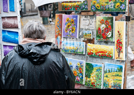 Exposition de peintures de rue de Montmartre Paris France // PARIS, France — Un visiteur observe une exposition extérieure de peintures, représentant des arrangements floraux et des représentations de la Tour Eiffel, d'un artiste de rue de Montmartre. Ce quartier emblématique parisien, en particulier la place du Tertre, est réputé pour sa communauté dynamique d’artistes de rue et son patrimoine bohème. Le charme historique et l'héritage artistique de Montmartre en font une destination populaire pour les touristes et les amateurs d'art. La région est connue pour ses chemins pavés et ses bâtiments historiques, contribuant à son caractère unique. Situé sur a la Banque D'Images