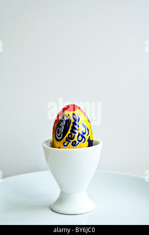 Un Cadbury Creme Egg in a white egg cup sur une plaque blanche avec fond uni. Banque D'Images