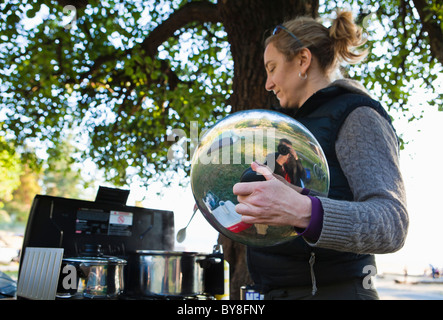 Un homme de prendre une photo reflète dans la casserole couvercle tenu par une femme la cuisson à l'extérieur sous un arbre. Banque D'Images