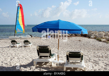Chaises vides, un parapluie bleu et un Hobie Cat sur la plage. Banque D'Images