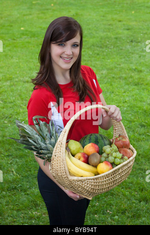 Jeune femme portant des vêtements de sport, souriant et tenant un panier de fruits Banque D'Images